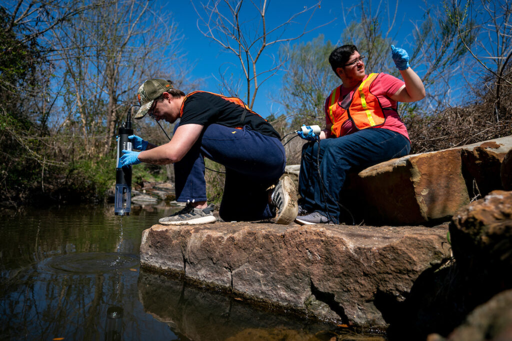 Two students use a meter to monitor water quality along a stream.