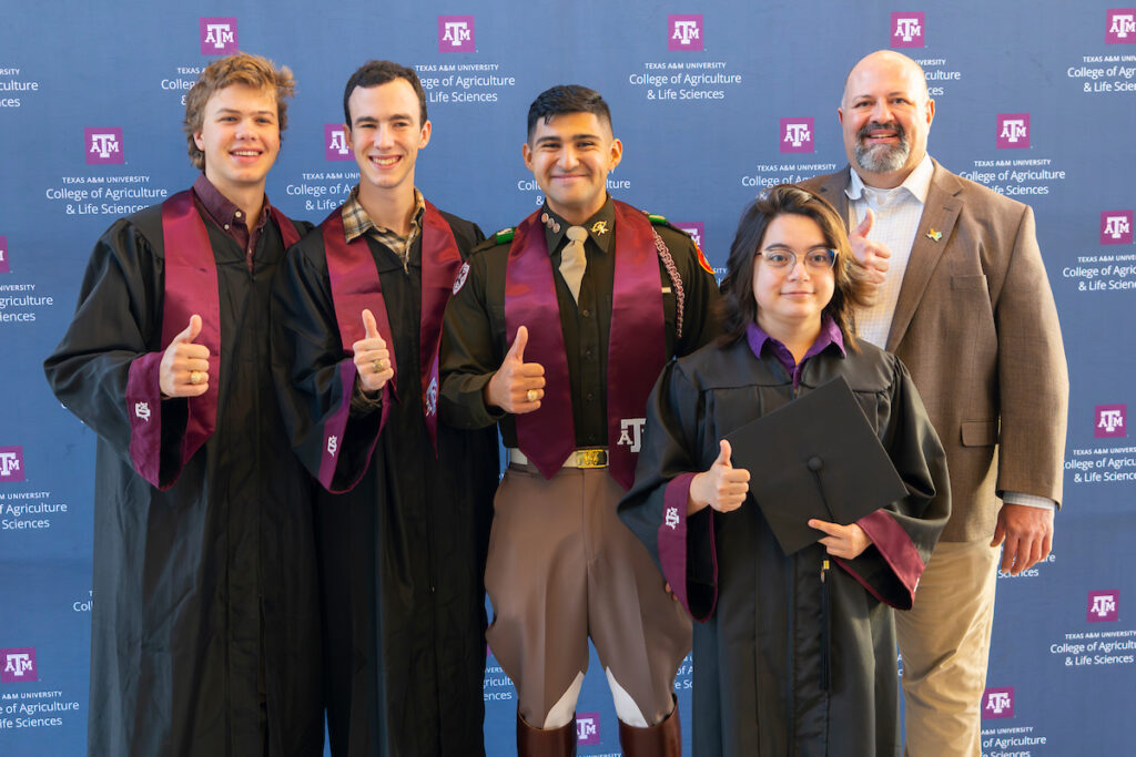 Four graduating students give a thumbs up. A man in a suit jacket standing next to them does the same. 