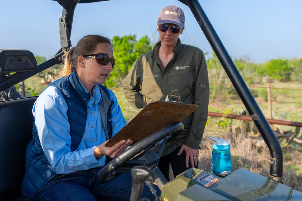 A woman on an all-terrain vehicle holds a clip board and speaks to another woman standing. 