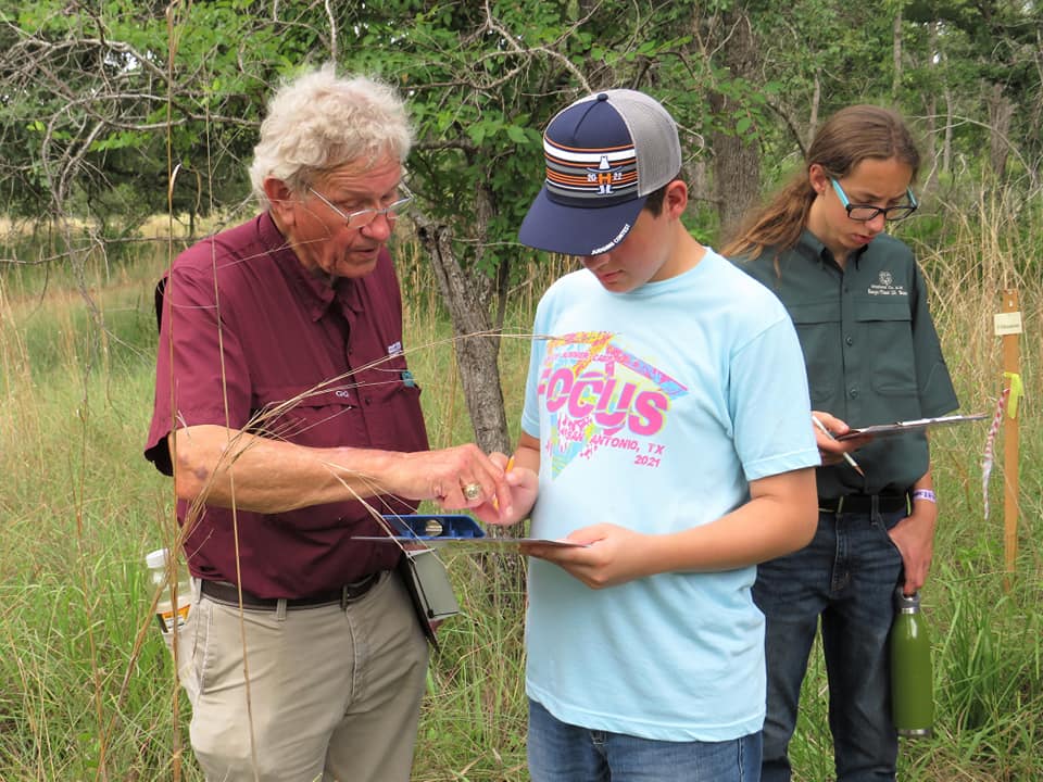 A man in a maroon shirt and glasses points to a clipboard being held by a young man in a cap and t-shirt. 