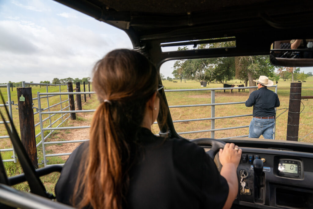 A woman driving an ATV waits for a man in a cowboy hat to open a gate to a cattle field. 