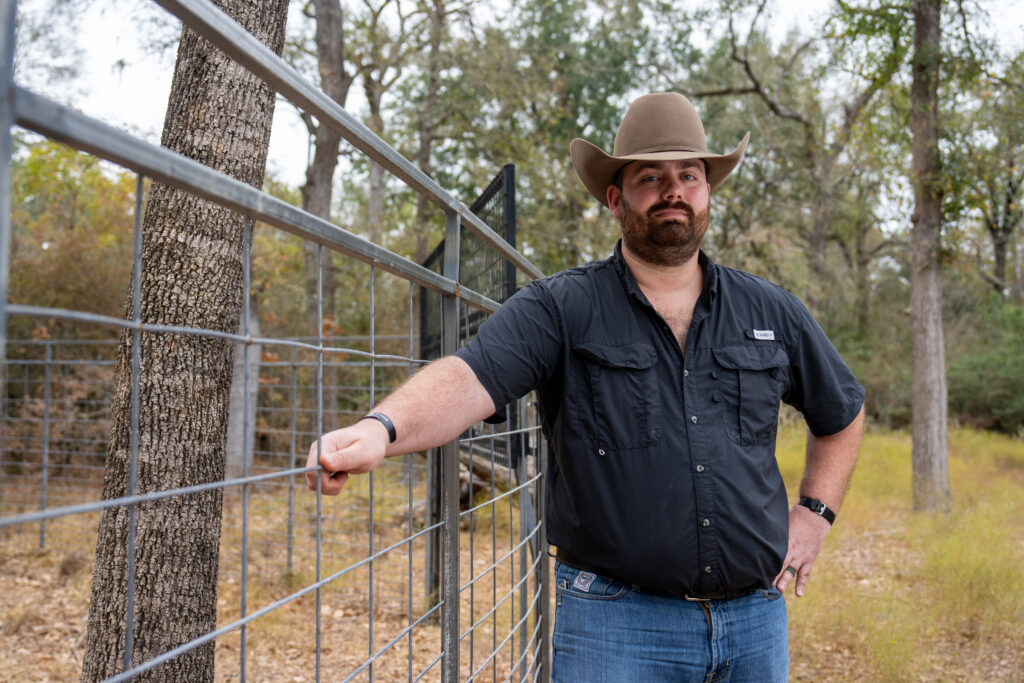 A man leans on a wire fence in the woods. He is wearing a dark blue short and jeans and a brown cowboy hat.