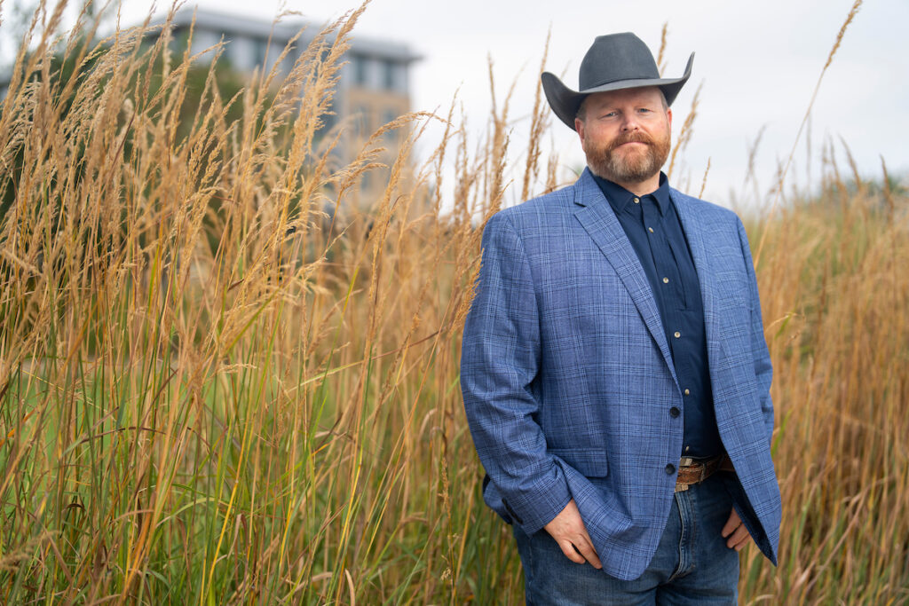 Jeff Goodwin stands in front of tall grass on the Texas A&M campus. He is wearing a blue jacket with a dark blue shirt and a gray cowboy hat.