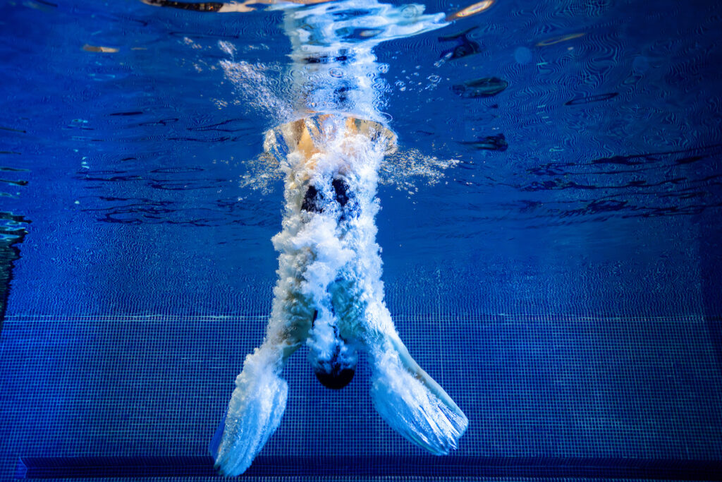 A diver is captured underwater, submerged headfirst with legs above, creating bubbles in a swimming pool