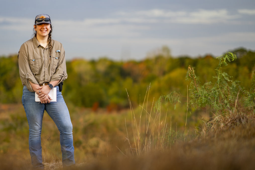 Cassandra Gomez standing in a tall grass field with a notebook in hand, as an example to students who are thinking about applying for admissions.