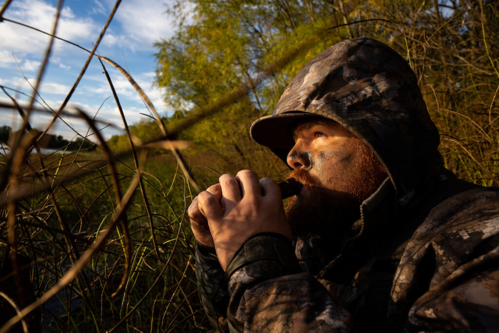 A man wearing camoflauge uses a duck call.