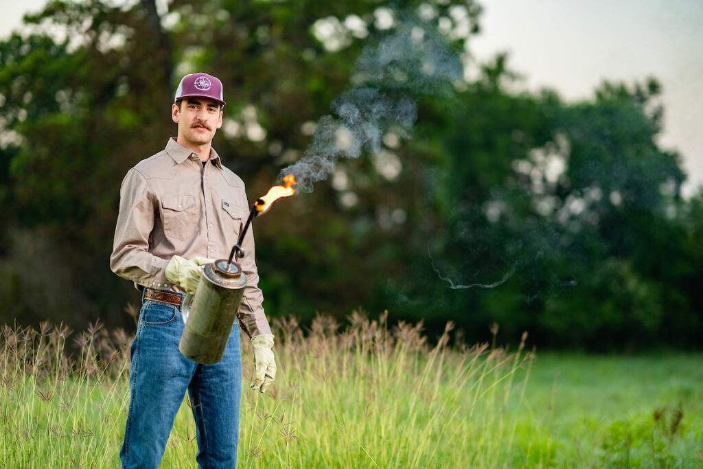 A man stands in a field holding a lit drip torch.