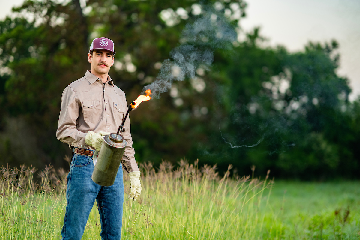 A man stands in a field holding a lit drip torch.