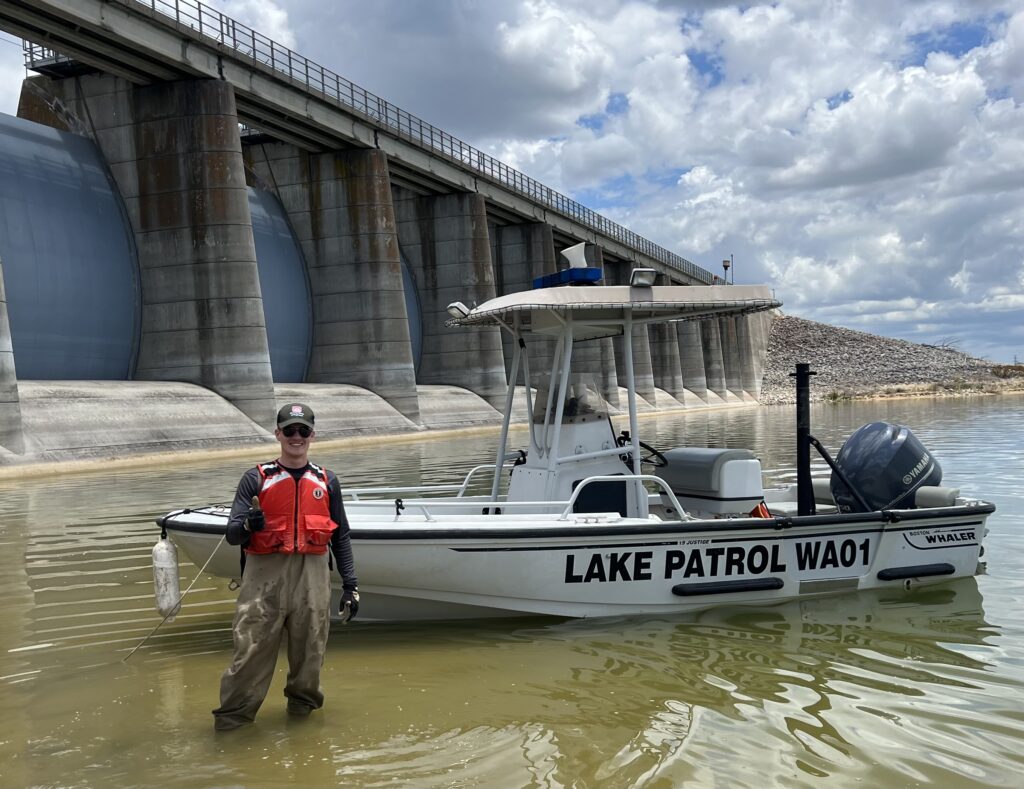 Man standing in shallow water with a high visibility vest on next to a boat that says "Lake Patrol WA01"