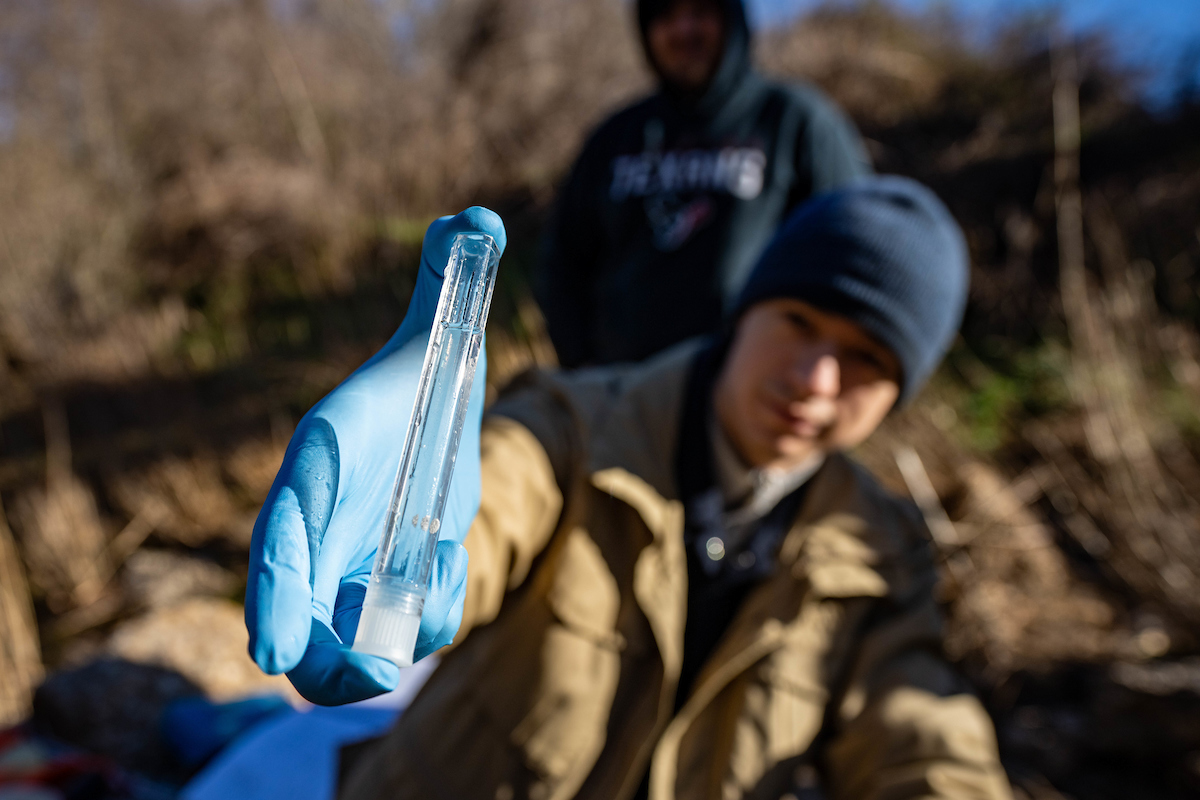 A young man extends a gloved hand holding a test tube of water.