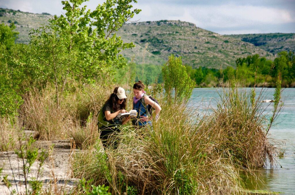 Two women look through a book next to a river.