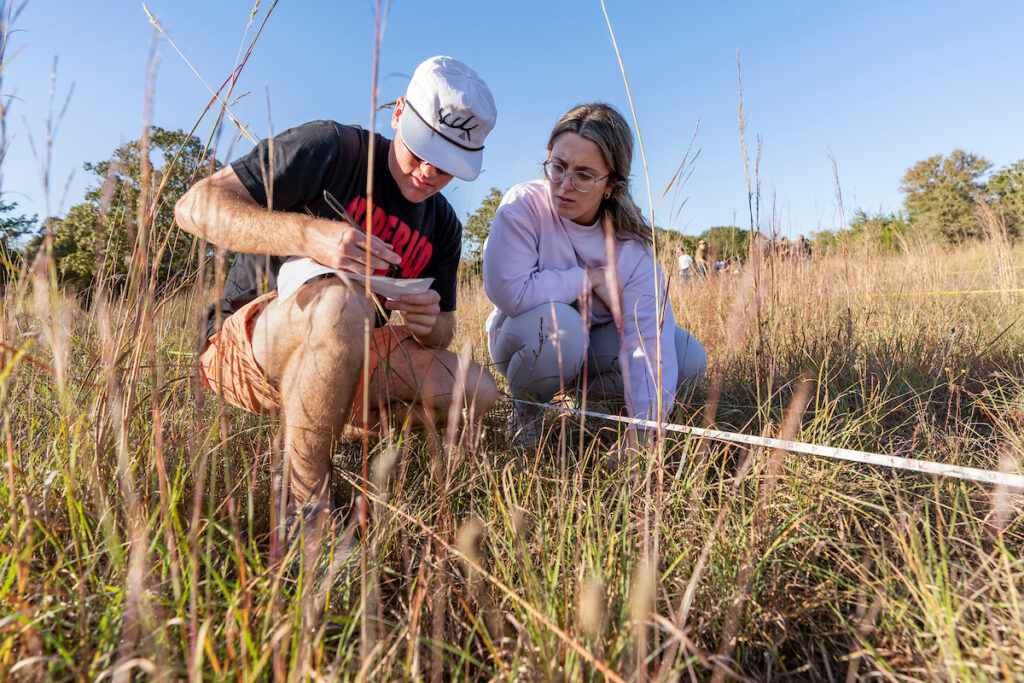 Two students crouching in tall grass. One is holding a pen and paper while the other watches
