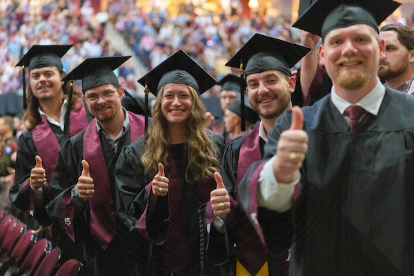 A group of five students at graduation with a cap and gown all holding up a Gig"Em thumbs up.